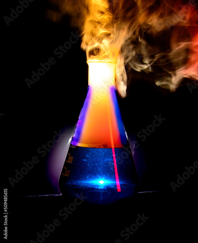 Strong chemical reaction with a lot smoke and vapors inside Erlenmeyer flask. Ignition is starting. Vessel with a blue liquid is standing on the table.  photo
