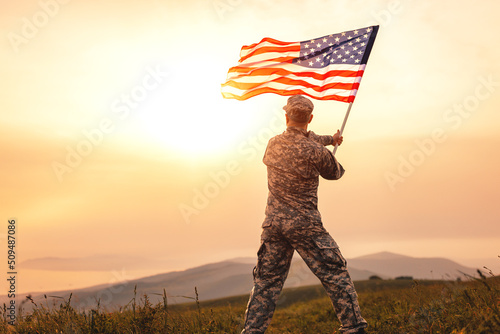 Male soldier in the uniform of the American army waving the US flag on top of a mountain in a clearing at sunset
