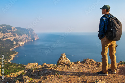 An adult man with photographic equipment stands on the edge of a cliff. Below is a picturesque sea bay with fishing boats.