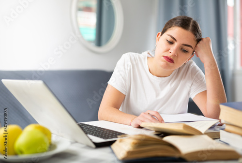 Photography Young tired and dissatisfied girl, doing lessons in a room on a laptop with books, reads a paragraph in a textbook, sitting