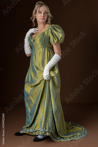 A blonde Regency woman wearing a green shot silk dress and standing against a plain backdrop