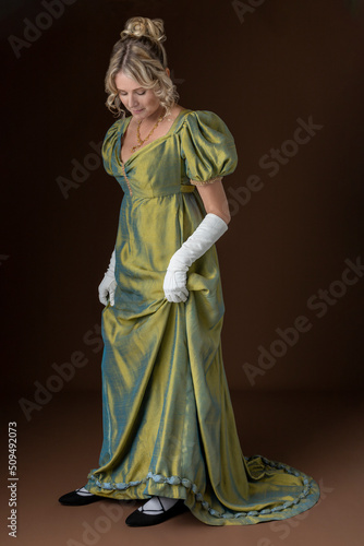 A blonde Regency woman wearing a green shot silk dress and standing against a plain backdrop