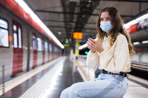 Girl wearing a protective mask, sitting on a subway platform bench during a pandemic, texts with friends on her mobile ..phone while waiting for a train