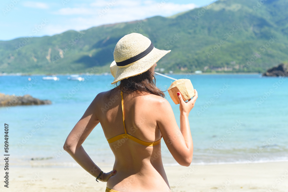 Young woman in yellow swimsuit drinking coconut on a sandy beach
