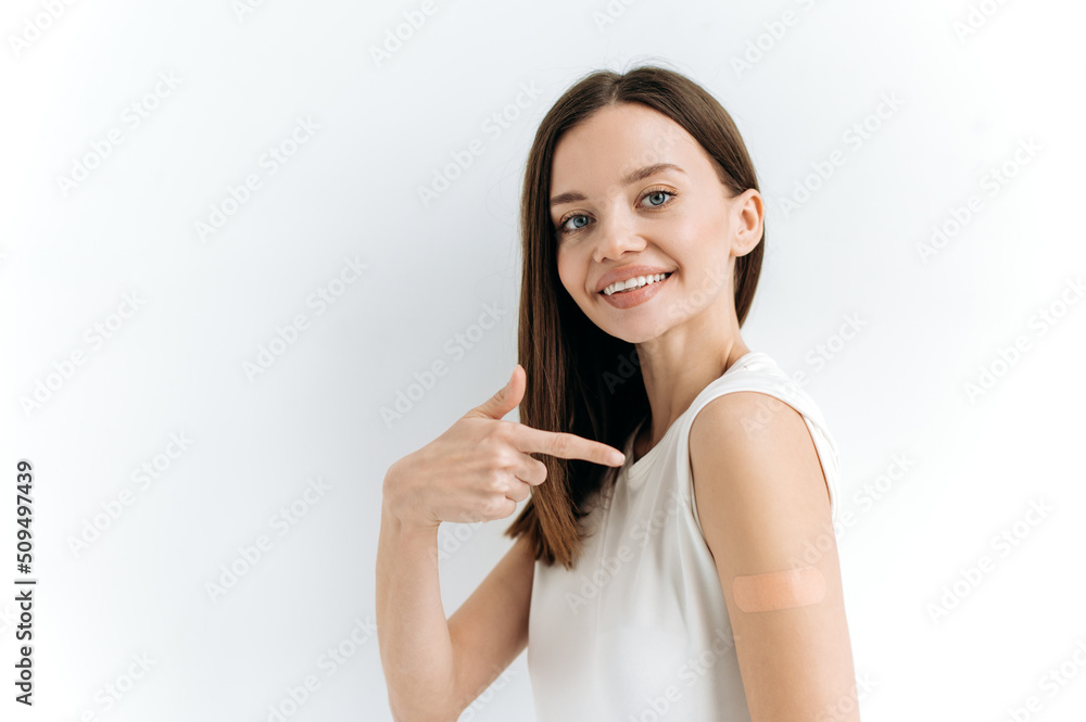Caucasian lovely blue-eyed girl, stands on an isolated white background with a plaster on her shoulder, received a vaccine against various diseases, acquired immunity, looks at the camera, smiles Caucasian lovely blue-eyed girl, stands on an isolated white background with a plaster on her shoulder, received a vaccine against various diseases, acquired immunity, looks at the camera, smiles