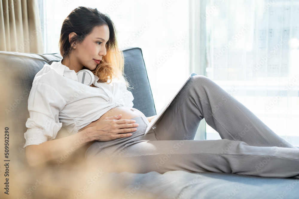 Asian young woman pregnant sitting and reading book with smile on sofa Asian young woman pregnant sitting and reading book with smile on sofa