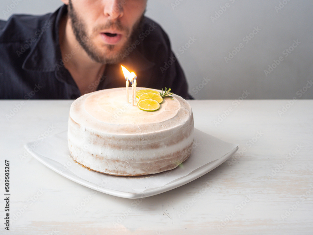 White Man Blows Candles on a Vegan Birthday Cake with Almond, Coconut and Lemon Base on a White Table and a White Background