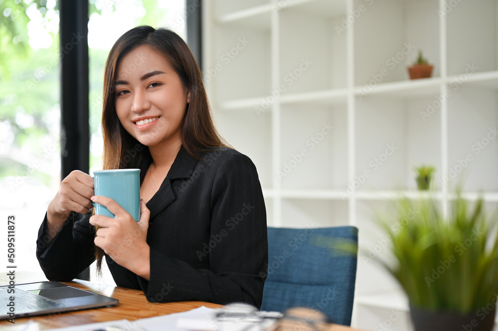 Asian businesswoman sits in office, holding a cup of coffee with positive smile expression. Asian businesswoman sits in office, holding a cup of coffee with positive smile expression.