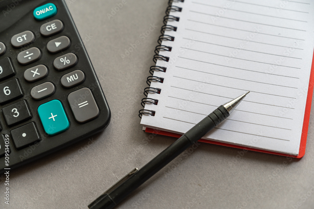 Calculator, pen, notepad lie on a gray surface. Open spring-loaded notebook with blank sheets. Close-up. Calculator, pen, notepad lie on a gray surface. Open spring-loaded notebook with blank sheets. Close-up.