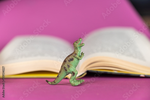 Dinosaur and open book against purple background. Green plastic miniature of an animal of prey. Hardcover book in the background. Selective focus.