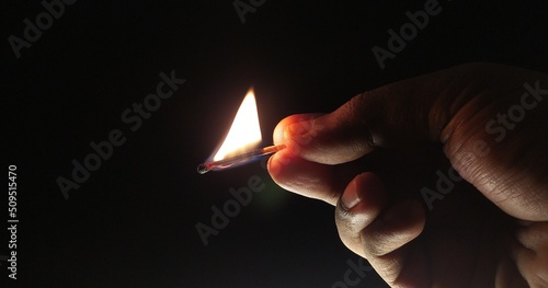 Man hand and finger holding one burning wooden matchstick with hot fire flame light isolated in a dark black room background with copy space. Close up macro side view. Power outage, darkness concept.
