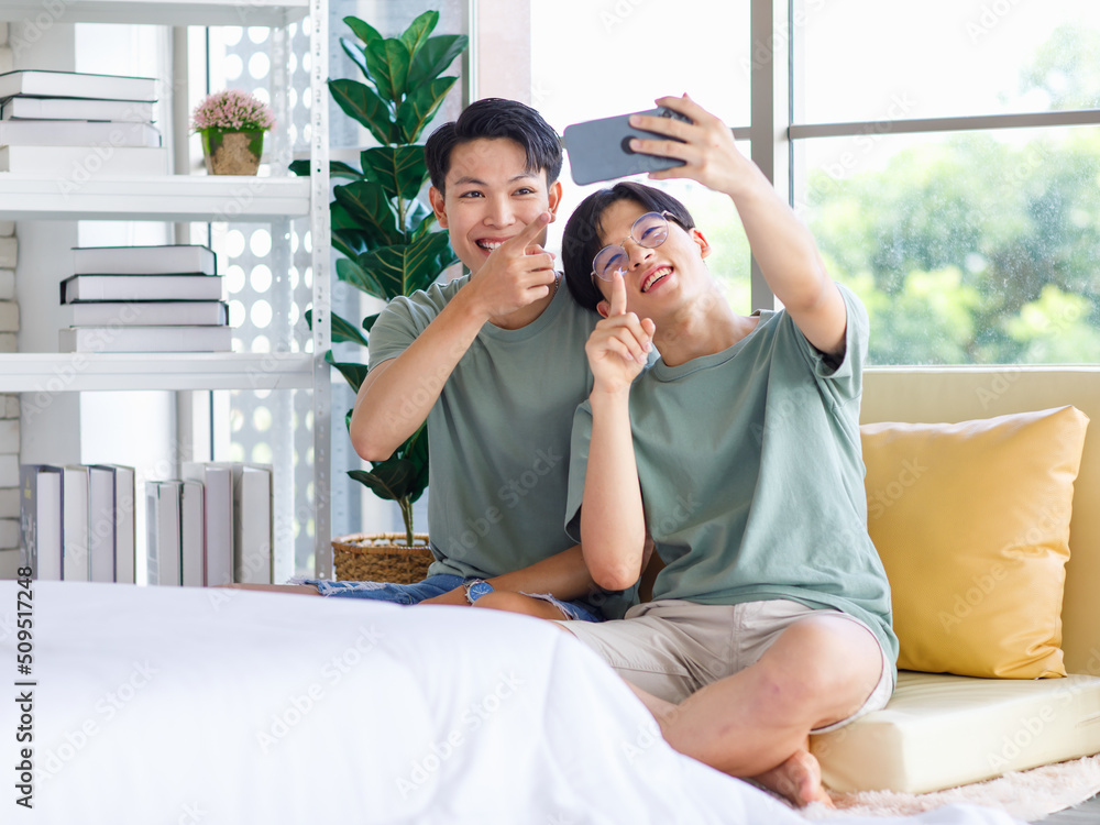 Two Asian young handsome pride male gay men lover couple partner sitting smiling together on cozy sofa in bedroom holding looking browsing surfing via smartphone cuddling hugging showing LGBTQ love