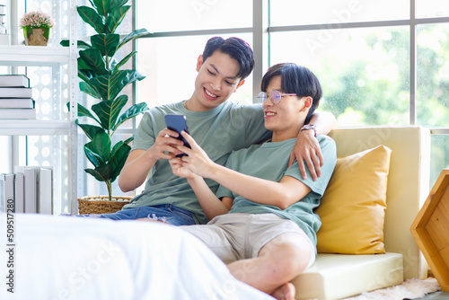Two Asian young handsome pride male gay men lover couple partner sitting smiling together on cozy sofa in bedroom holding looking browsing surfing via smartphone cuddling hugging showing LGBTQ love