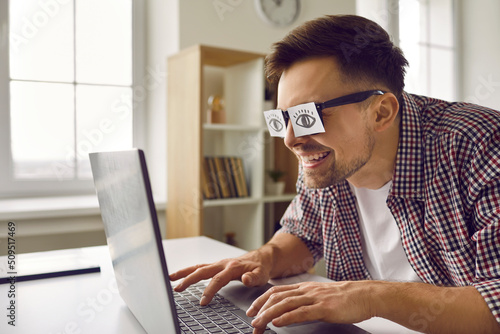 Funny weird sleepy man with sticker eyes glued to eyeglasses working on modern laptop. Tired university student with sticky pieces of paper on his eyes pretending to be studying on notebook computer photo