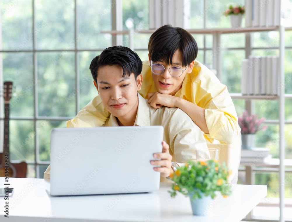 Two Asian young happy pride teenager male gay men lover couple partner sitting smiling hugging cuddling together in front laptop notebook computer browsing surfing using credit card shopping online