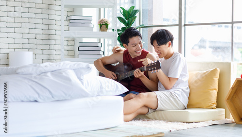 Two Asian young happy romantic lovely teenager male gay men lover couple partner sitting smiling on cozy sofa in bedroom cuddling hugging together while husband playing acoustic guitar and sing song