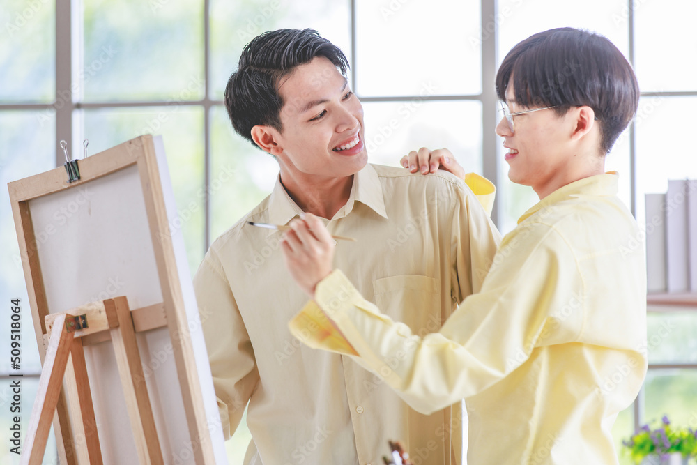 Two Asian young lovely pride teenager male gay men lover couple partner standing smiling having fun playing paintbrush in front canvas painting board on easel in glass windows workshop studio at home Two Asian young lovely pride teenager male gay men lover couple partner standing smiling having fun playing paintbrush in front canvas painting board on easel in glass windows workshop studio at home