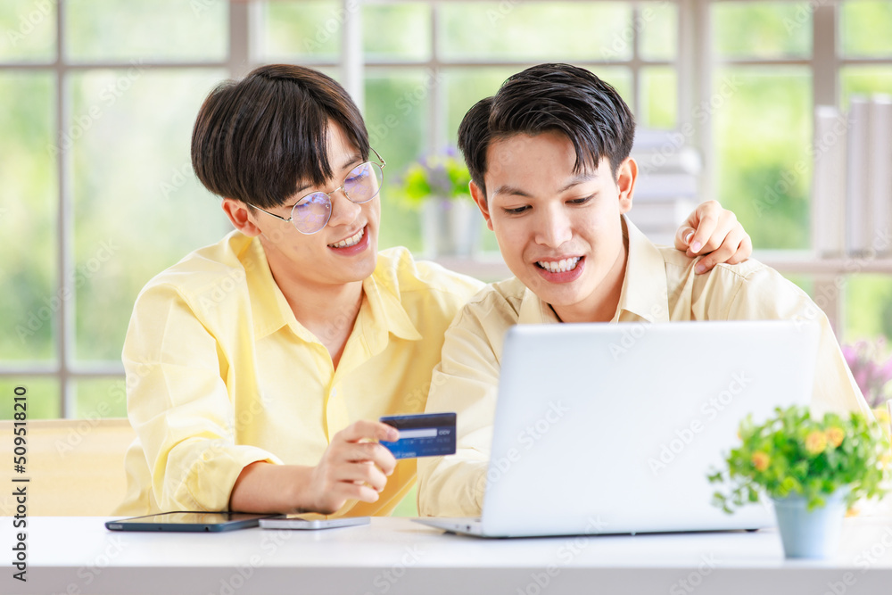 Two Asian young happy pride teenager male gay men lover couple partner sitting smiling hugging cuddling together in front laptop notebook computer browsing surfing using credit card shopping online Two Asian young happy pride teenager male gay men lover couple partner sitting smiling hugging cuddling together in front laptop notebook computer browsing surfing using credit card shopping online