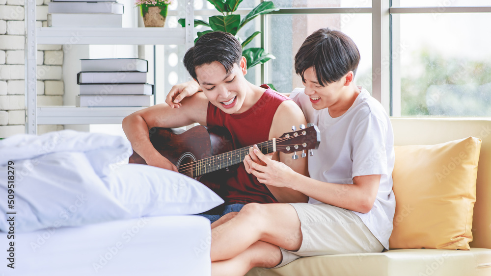Fototapeta premium Two Asian young happy romantic lovely teenager male gay men lover couple partner sitting smiling on cozy sofa in bedroom cuddling hugging together while husband playing acoustic guitar and sing song