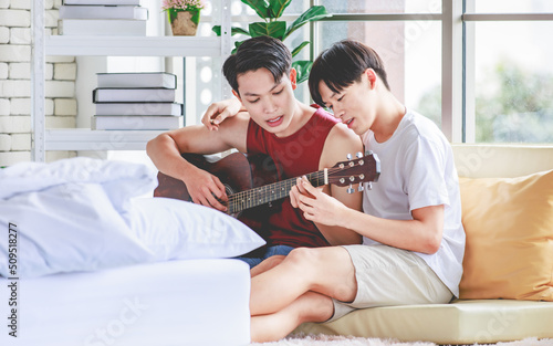 Two Asian young happy romantic lovely teenager male gay men lover couple partner sitting smiling on cozy sofa in bedroom cuddling hugging together while husband playing acoustic guitar and sing song