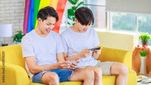 Two Asian cheerful teenager male gay men lover couple partner sitting smiling on sofa holding smartphone playing fun digital game online together in living room with rainbow pride equality unity flag