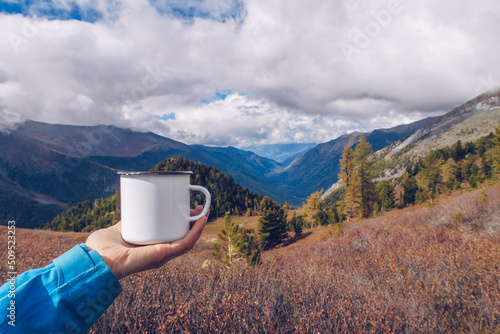 Hand holding enamel white mug mockup with forest and mountains valley background. Trekking merchandise and camping gear. Stock wildwood photo with white metal cup. Rustic product template.