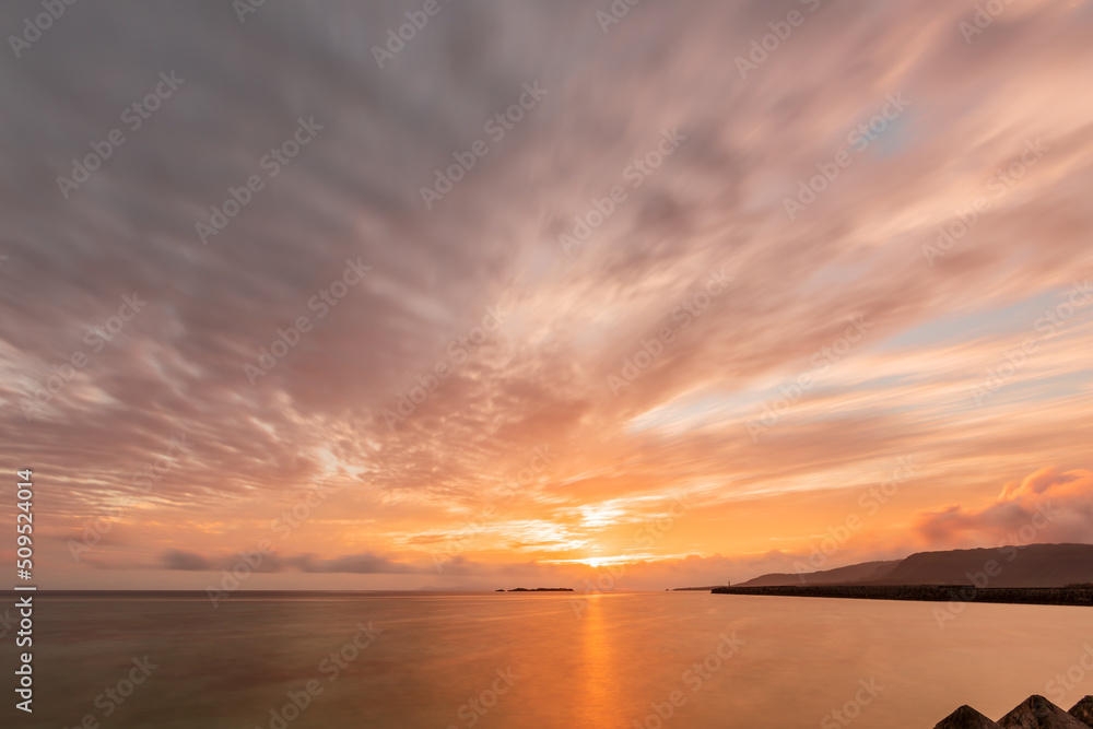 Gorgeous sunrise on horizon, colorful clouds in motion blur, sun reflected on sea surface.