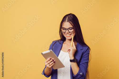 Adorable brunette in glasses with long hair dressed in dark blue suit cute smiling, holding her tablet, looks happy, makes video call, over yellow background with copy space. People sincere emotions.