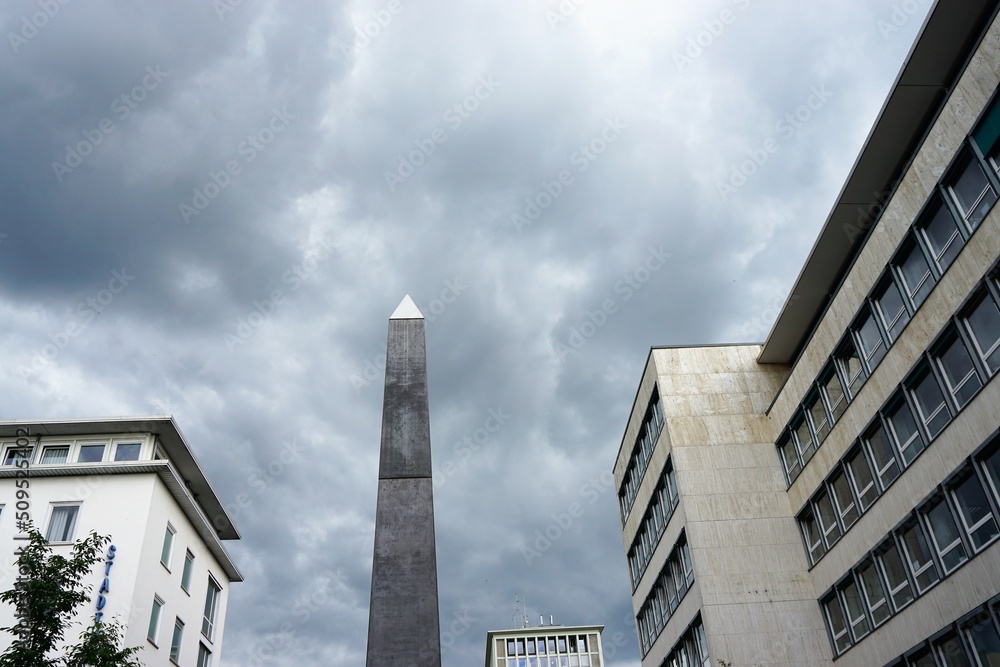 Weiße und graue Häusergebäude mit Obelisk-Statue in Mitte vor Himmel mit dunklen Wolken in Stadt am Nachmittag im Frühling 