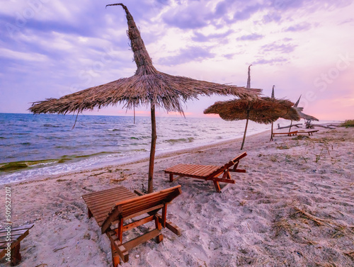 Beautiful sunrise or sunset over the calm waters of the Black Sea on the Kinburg Spit in Ukraine. Beach summer landscape. Straw umbrella and wooden deck chair on the beach at sunset or sunrise photo