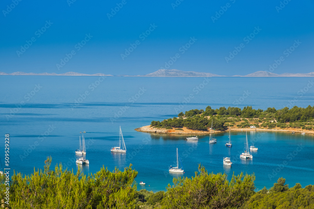 Obraz premium Panoramic view on Kosirina lagoon on Murter island in Croatia, anchored sailing boats and yachts on blue sea, Kornati archipelago in background