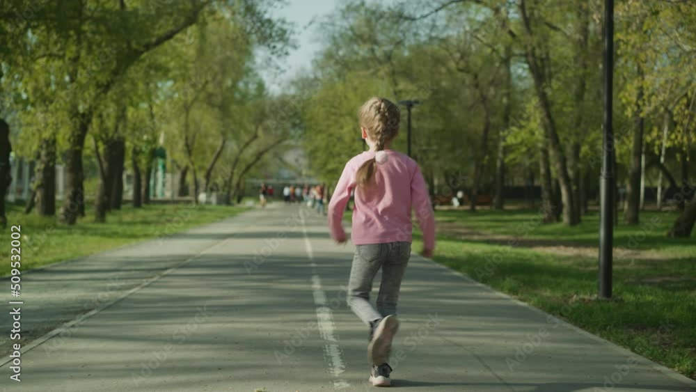 Playful little girl with braided fair hair runs along empty asphalt pedestrian road in spring park backside view. Active sportive child has fun slow motion Playful little girl with braided fair hair runs along empty asphalt pedestrian road in spring park backside view. Active sportive child has fun slow motion