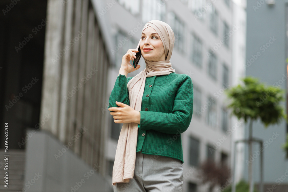 Muslim business woman in green jacket and hijab standing on street and talking on mobile. Confident female entrepreneur having working conversation on phone, outdoor on background of modern buildings