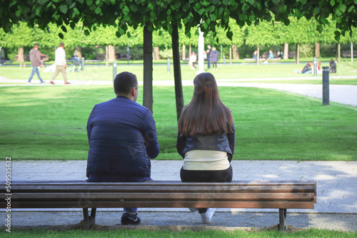 View from behind on a couple sitting on long bench keeping distance and talking in green summer park
