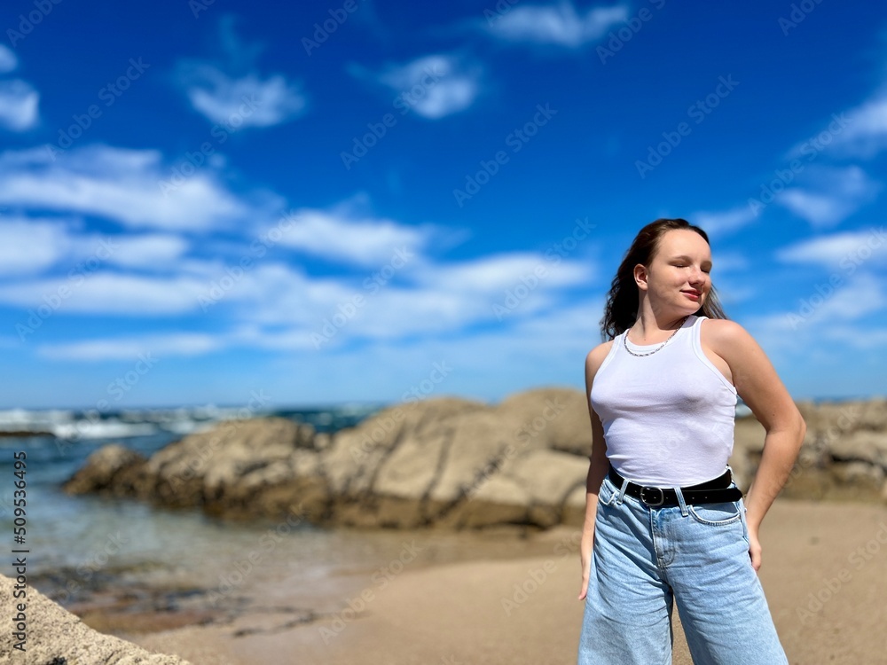 a teenage girl stands against the backdrop of the Atlantic Ocean in the city of Pante Lima, the water of the sea and large stones are visible, she looks back into the frame, she is thoughtful. a teenage girl stands against the backdrop of the Atlantic Ocean in the city of Pante Lima, the water of the sea and large stones are visible, she looks back into the frame, she is thoughtful.