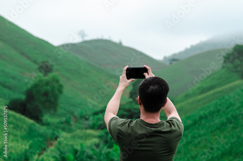 A male person using mobile phone taking a photo of greenery mountain when hiking with fog in the morning. © baramyou0708