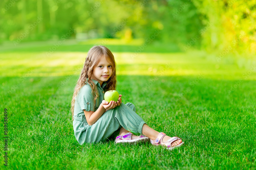 Fototapeta premium a happy child girl in the summer on the lawn with a green apple sits on the grass and smiles