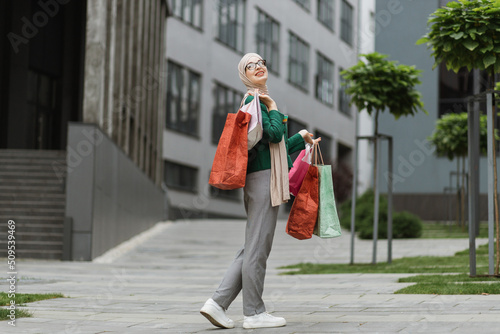 Pretty young muslim woman, wearing grey pants and green jacket, walking at the street with colorful shopping bags, smiling and looking outside. Shopping in the city concept. © sofiko14