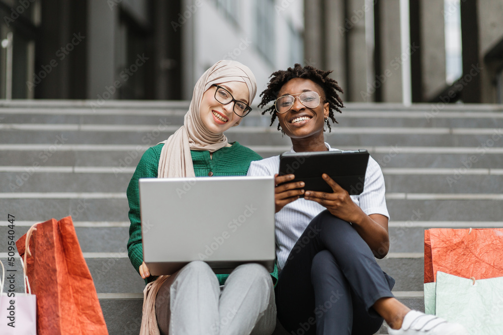 City shopping concept. Outdoor urban lifestyle portrait of two diverse young women, African and Muslim, sitting on the steps of the mall with a laptop and tablet and shopping online. City shopping concept. Outdoor urban lifestyle portrait of two diverse young women, African and Muslim, sitting on the steps of the mall with a laptop and tablet and shopping online.