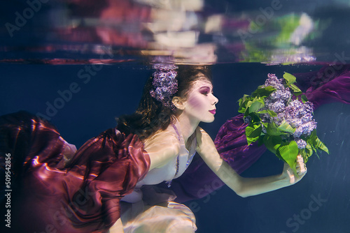 woman with lilac bouquet in colorful clothes on the dark background swimming underwater 