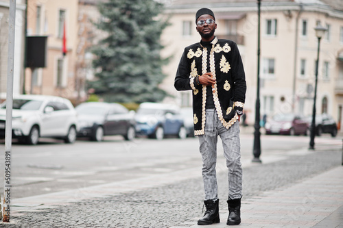 Mega stylish african man in traditional jacket pose. Fashionable black guy in hat and sunglasses with cigar in hand.