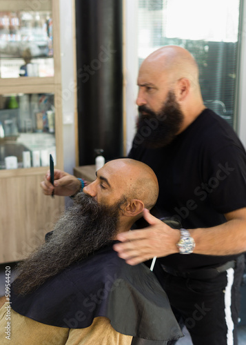 Bald bearded man sitting on a client seat being served by a male barber into his place of work