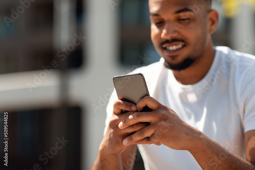 Closeup shot of african american man using cellphone, texting or surfing internet, sitting outdoors, focus on device