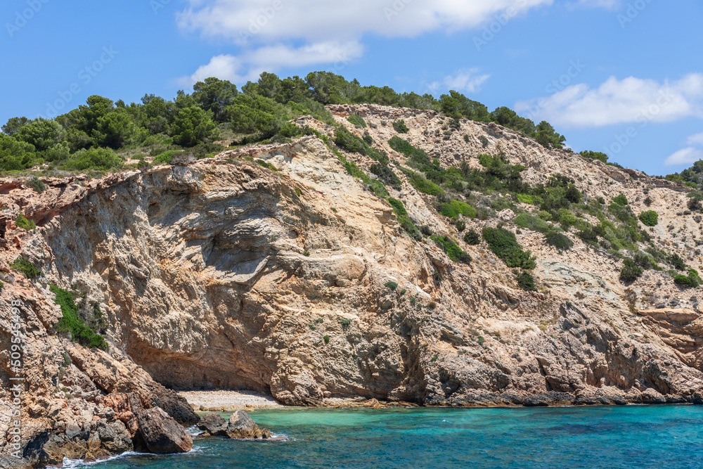 Part of steep coast, layers of centuries-old rock are clearly visible, covered with green bushes and moss, clear sea, Ibiza, Balearic Islands, Spain