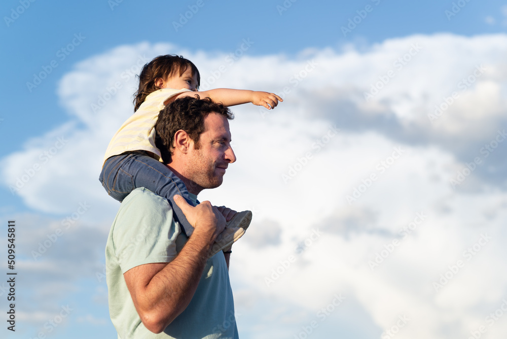 Side view of a father carrying her little girl on shoulders while she is pointing out forwards on a background of blue sky with clouds.