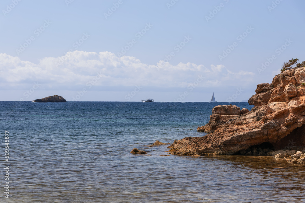 Centuries-old tandem of bright yellow sandy rock and blue calm sea with ripples, sailing and motor yachts on horizon. Ibiza, Balearic Islands, Spain
