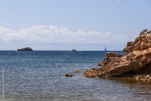 Centuries-old tandem of bright yellow sandy rock and blue calm sea with ripples, sailing and motor yachts on horizon. Ibiza, Balearic Islands, Spain