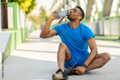 Handsome African American guy in sportswear drinks a bottle of water or a sports drink, sits on the floor, empty space. photo