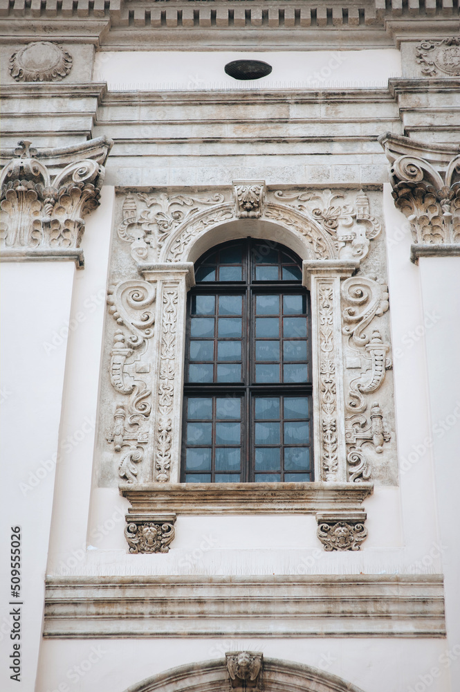 Antique openwork baroque window with a lattice frame and decorative stucco on a light cream wall. Jesuit Church in Lviv. Antique openwork baroque window with a lattice frame and decorative stucco on a light cream wall. Jesuit Church in Lviv.