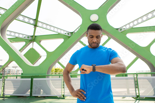  African man in sportswear exercising with a fitness bracelet, blurred background on a bridge